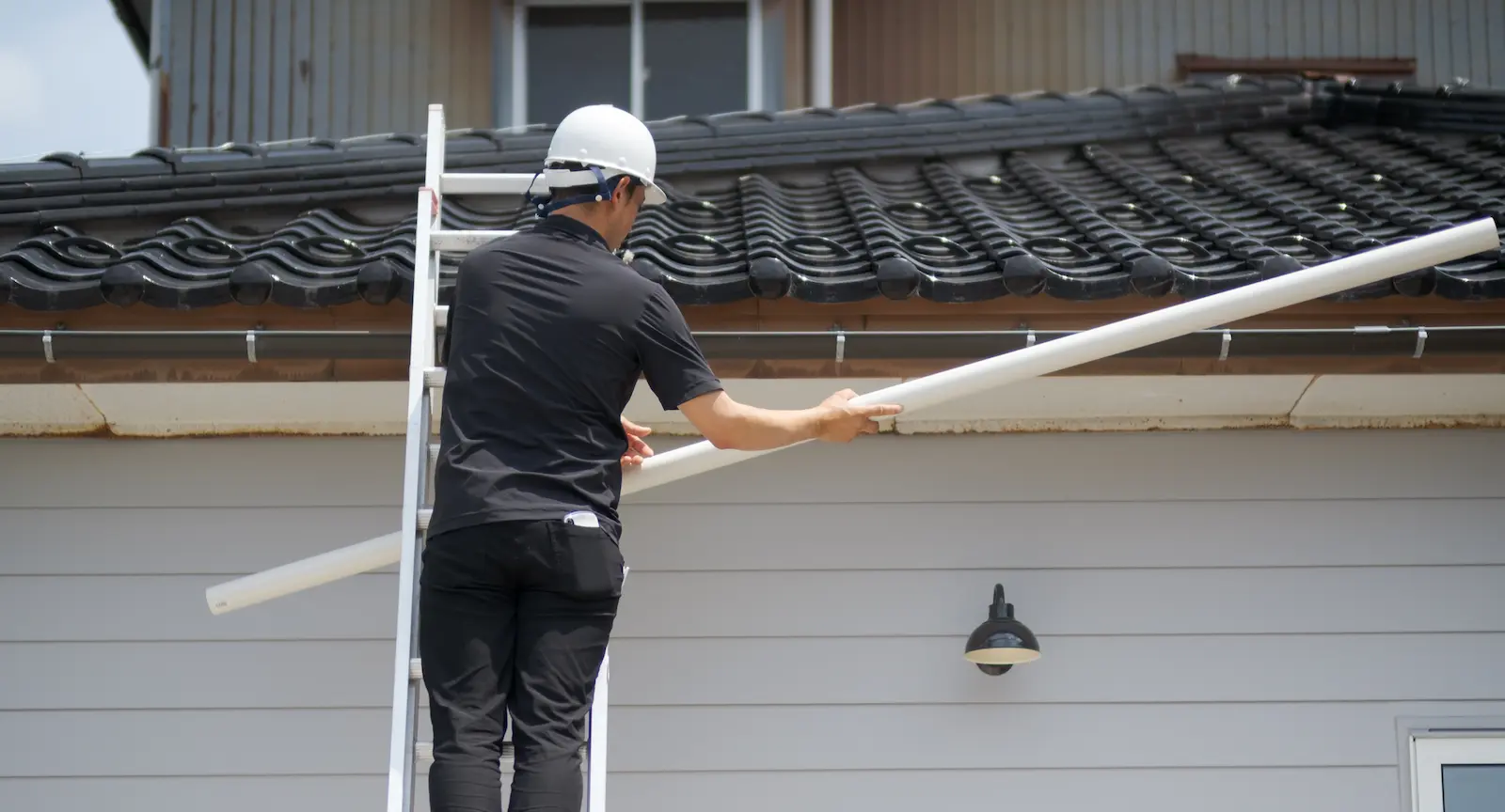 梯子を使用して屋根上の現場へ雨樋の交換部材を搬入する職人