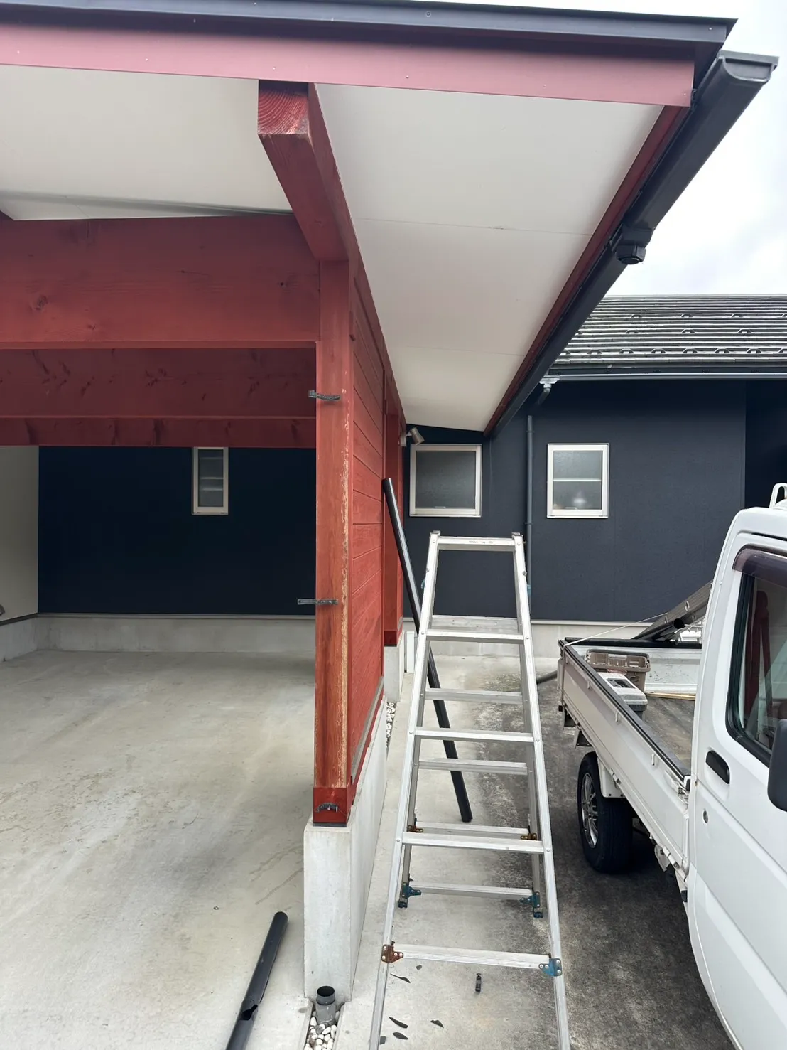 車庫の赤い柱に取り付けられた雨樋の交換工事を行うためにはしごとトラックを配置した施工前の様子
